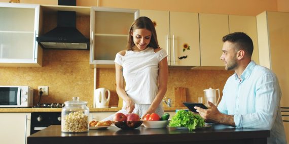 Attractive couple chatting in the kitchen early morning. Handsome man using tablet while his girlfriend cooking breakfast at home